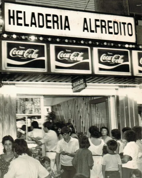 Foto antigua en blanco y negro del local: cartel «Heladería Alfredito», carteles de Coca-Cola y mucha gente en la puerta
